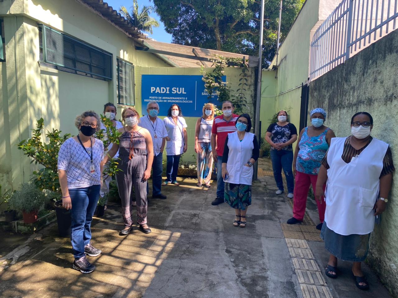 Diante de um prédio amarelo, sede do Padis Sul 11 pessoas todas de máscara, posam respeitando o distanciamento, elas alternam os espaços, são 9 mulheres e dois homens, seis profissionais estão uniformizados com avental